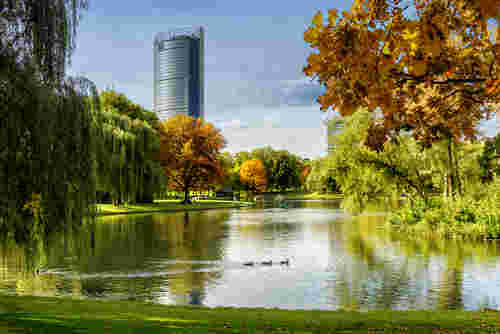 Städtischer Park mit Teich, buntem Herbstlaub und schwimmenden Enten, im Hintergrund ein moderner Hochhausturm vor blauem Himmel. Städtischer Park mit Teich, buntem Herbstlaub und schwimmenden Enten, im Hintergrund ein moderner Hochhausturm vor blauem Himmel.